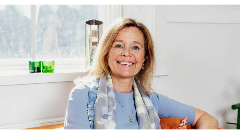 Asa wearing a light blue dress with scarf sitting on a sofa, and smiling.